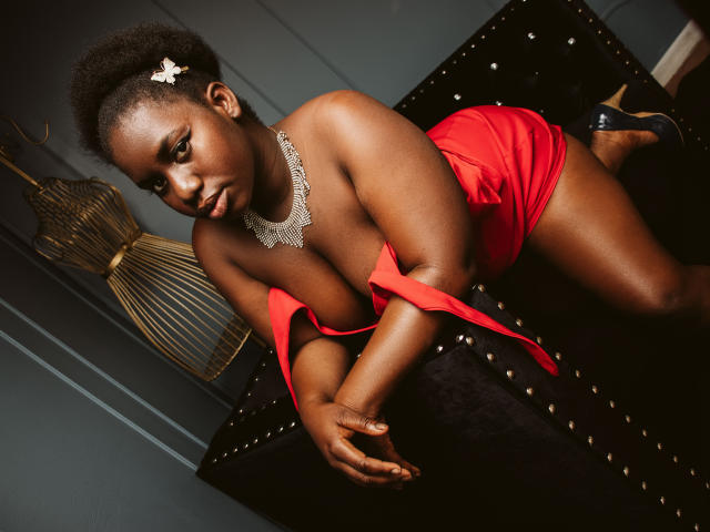 Dilay with dark skin, wearing a red dress, showing her big ass and tits, leaning on a black studded couch, black heels, silver necklace, and a white hair clip.