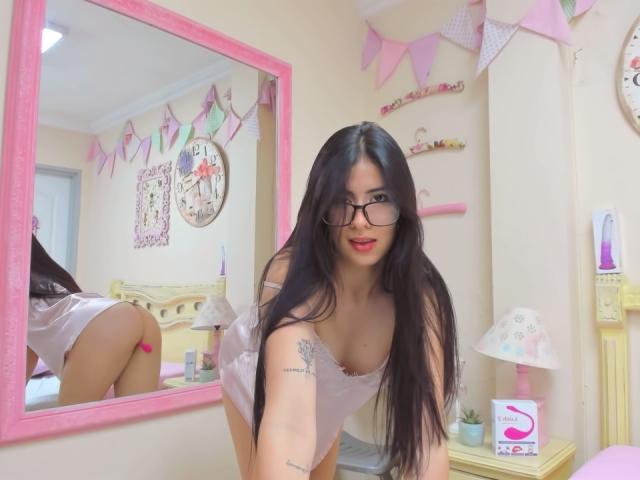 Sarah Rogerss, avec ses longs cheveux noirs, ses lunettes et ses lèvres roses, est penchée dans une pièce rose, vêtue d'une chemise de nuit blanche, montrant ses fesses et un tatouage sur son bras droit..