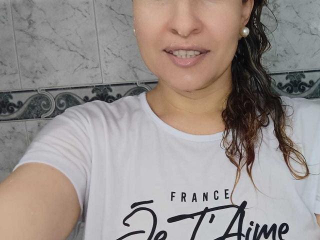 EroticSasha, wet brunette hair, white shirt, pearl earrings, smiling, visible nose ring, slightly chubby face, soft skin, subtle makeup, slightly messy shirt, selfie, upper body, casual.