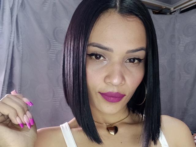 Lily Clark tiene un corte de pelo bob negro y elegante, lápiz labial morado y uñas rosas. Lleva una camiseta blanca y un collar con forma de corazón dorado, además de aros grandes..