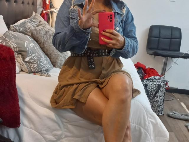 AmberDemons sits on a bed, wearing a denim jacket and brown dress with a belt, holding a red phone, showing off her thighs and crossed legs, with her tits visible.