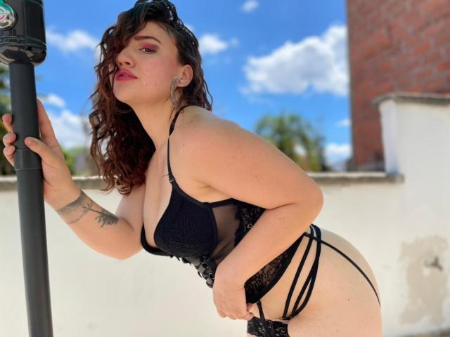 AgnesFortune, avec ses cheveux bruns bouclés et son rouge à lèvres rose, est appuyée contre un poteau, vêtue de lingerie noire en dentelle, montrant ses seins et ses fesses, avec un tatouage sur son bras gauche, sous un ciel bleu..