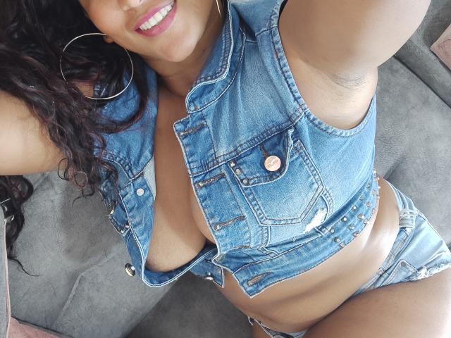 YiyaHot69 with curly black hair, smiling, wearing a blue denim vest, exposing large tits, and ripped denim shorts, sitting on a grey couch, showing armpit.