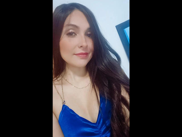 AmbeerBlack has long brown hair, blue dress with a deep V, small tits, subtle makeup, and a slight smile. She's wearing a gold necklace, looking cute.