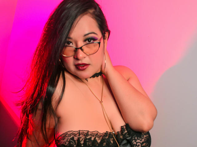 AbriilWilliamis tiene el pelo largo y negro, usa gafas y una blusa negra de encaje, mostrando sus pechos con un collar dorado, con una iluminación rosa y roja. Está tocándose la cara..