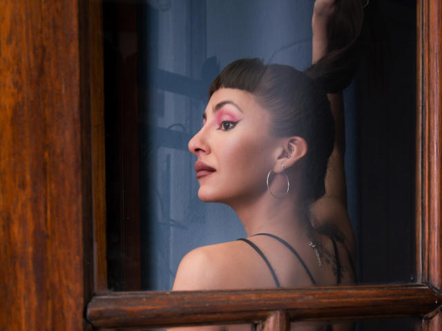 Eliza Wells, vue de profil, avec ses cheveux bruns attachés en chignon, portant un maquillage rose, des boucles d'oreilles pendantes et un haut noir à bretelles spaghetti, debout près d'une fenêtre en bois..