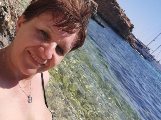DoraBlueEyess with short brown hair, smiling, wearing a black tank top and silver necklace, standing by clear, shallow water. She has small, cute tits and a cute ass.