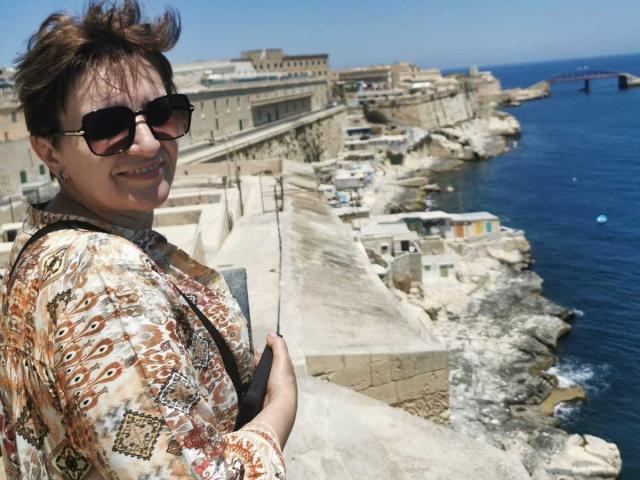 DoraBlueEyess with short brown hair, wearing sunglasses and a patterned shirt, stands on a rocky cliff, holding a camera, smiling at the blue ocean.