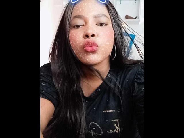 Emira, mit langen schwarzen Haaren, trägt ein schwarzes Hemd und hat glitzerndes Puder im Gesicht, volle, rote Lippen und Reifenohrringe. Sie macht ein Küsschen-Gesicht in einem Selfie..