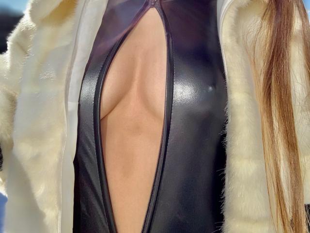 La top nera in pelle con inserti a vista di LuxuryMinyDiva rivela il suo seno, con un giubbotto di pelliccia bianca aperto, che lascia intravedere il suo petto e una piccola parte della sua pelle abbronzata. Lunghi capelli castani..