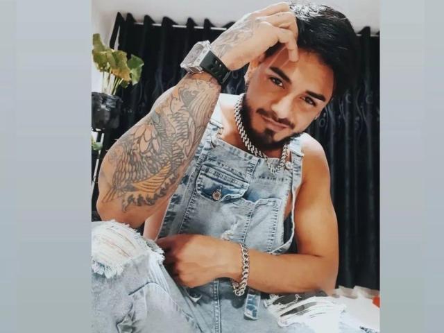 Homme avec des tatouages sur le bras droit, portant une salopette en jean déchirée, une montre noire et un collier en chaîne, assis, une main sur la tête, souriant, avec des cheveux courts et foncés, et une barbe, dans une pose décontractée..