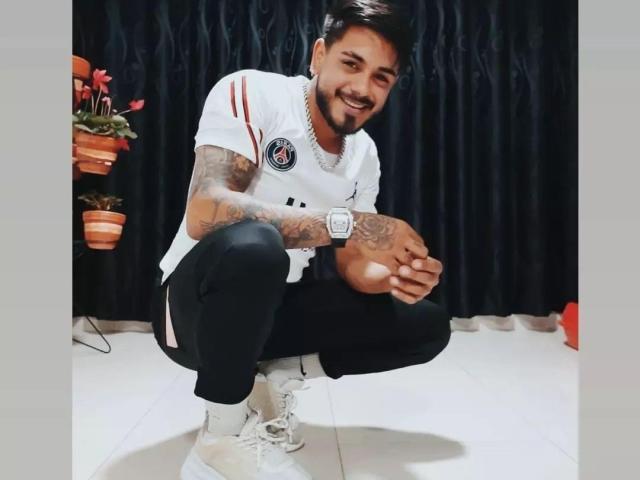 VisiI, hockend und lächelnd, hat Tattoos auf seinen Armen, trägt ein weißes Hemd, schwarze Hosen, weiße Turnschuhe und eine Uhr, vor einem dunklen Vorhang..
