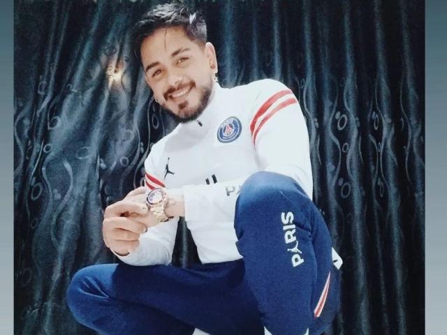 Un homme, avec des cheveux noirs courts, une barbe et un sourire, accroupi, portant une veste blanche Puma avec des rayures rouges, un pantalon bleu avec le mot "Puma" sur le côté, tenant une montre..