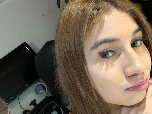 SaraPleasure's face with light brown hair, green eyes, and pink lips. She has black eyeliner, thin eyebrows, and a slight smile. Her nose is visible, and she's looking at the camera.