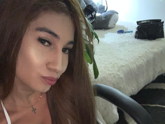Sara a de longs cheveux bruns, des sourcils épais et du rouge à lèvres rose. Elle a un petit sourire en plissant les lèvres, elle porte un haut blanc et un collier avec une petite croix..