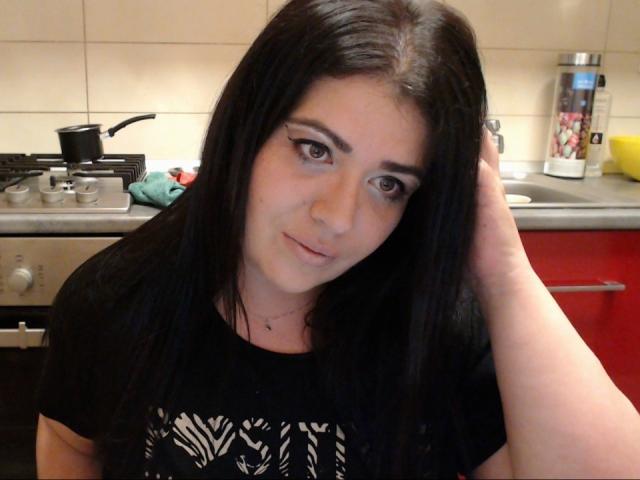 Carolina69 with long black hair, wearing a black shirt with a zebra pattern, hand in hair, looking at camera, kitchen background with stove and red cabinet.