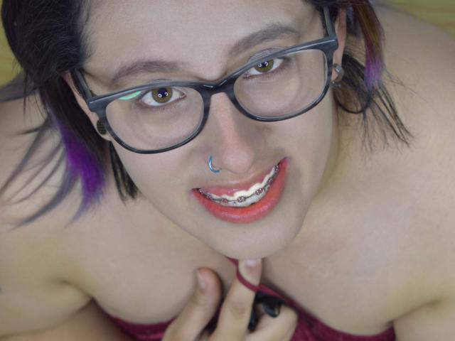 NatalieHarris with glasses, nose ring, and purple hair, smiling with teeth showing, touching her cleavage, showing her tits, wearing red lipstick, and black ear gauges.