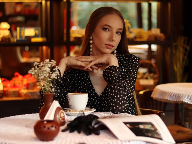 SafiraFuracao with long brown hair, wearing a black polka-dot blouse, pearl earrings, and black gloves, resting her chin on her hands, with a white cup and small vase.