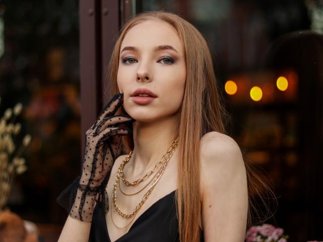 SafiraFuracao with long brown hair, wearing a black dress, gold chains, and sheer polka-dot gloves, looking sultry with her hand near her face. Her tits are small.
