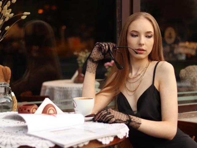 SafiraFuracao with long blonde hair, black lace gloves, black dress, gold necklace, holding eyeglasses, sitting at a table with a cup, magazine, and lace cloth.