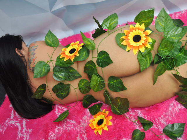 GiorginaMara, nuda, sdraiata su un tappeto rosa e soffice, con la schiena rivolta verso la fotocamera, con girasoli e foglie verdi che le coprono il sedere, lunghi capelli neri, tatuaggio sulla parte superiore della schiena, pelle abbronzata..