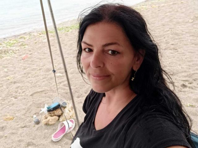 YulliaMilfX with dark hair, brown eyes, small tits, wearing black shirt, earrings, standing on beach, holding fishing pole, pink sandals on sand, water and grass in background.