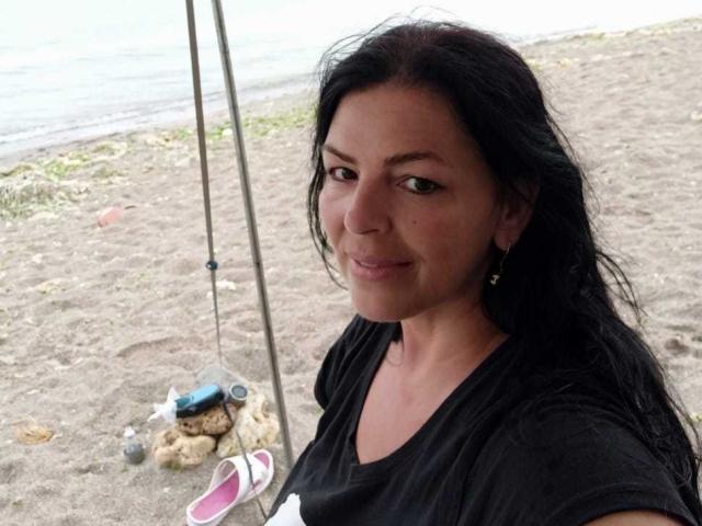 YulliaMilfX has dark, wavy hair and is smiling slightly, wearing a black shirt, holding fishing poles with pink shoes in the sand, near a water body.