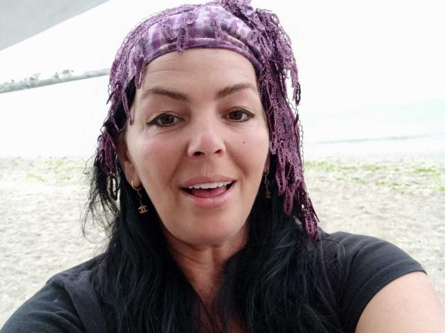 YulliaMilfX with purple, dreads-like headwrap, black hair, brown eyes, small nose, smiling, wearing black shirt, gold earrings, soft skin, slight tan, medium breast size.
