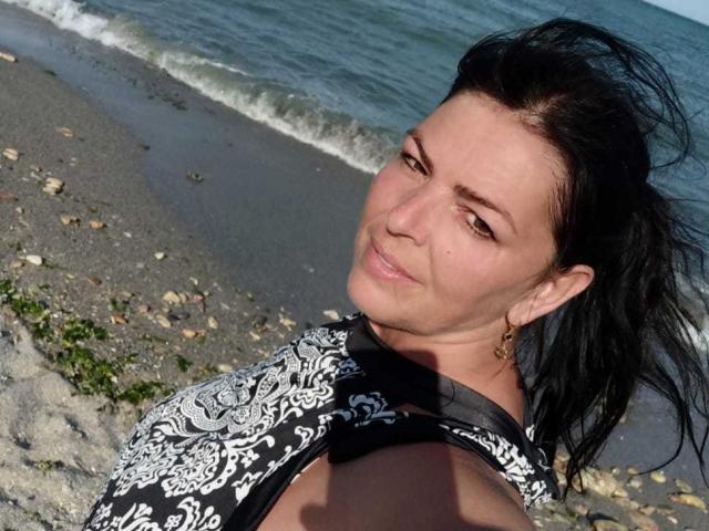 YulliaMilfX with black hair, brown eyes, and small gold earrings, wearing a black and white patterned top, smiling at the camera on a rocky beach with gentle waves.