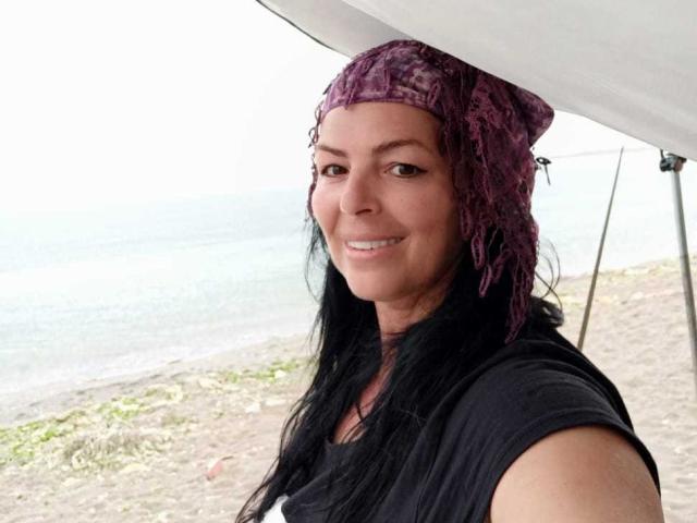 YulliaMilfX with dark hair, purple headscarf, black shirt, smiling, standing near a beach, holding a white umbrella, visible ocean in background, cute, attractive.