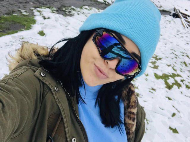 NicoColeman with black hair, blue beanie, reflective sunglasses, green jacket with fur trim, and blue shirt, standing in snowy, grassy area. Boobs not visible.