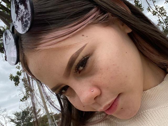 Jazmin Wells hat helle Haut, dunkelbraue Augenbrauen, langes, braunes Haar mit rosafarbenen Spitzen und ein Nasenpiercing. Sie trägt einen beigen Pullover und eine Sonnenbrille auf dem Kopf..