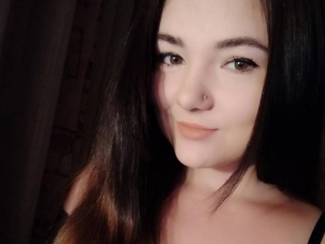 Uma pessoa de etnia não especificada, com pele clara, olhos castanhos, sobrancelhas pretas, piercing no nariz e cabelo castanho longo, sorrindo discretamente, vestindo uma blusa preta, olhando para a câmera..