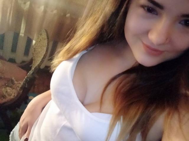Uma mulher com cabelo castanho claro, sorrindo, vestindo uma camiseta branca que mostra seus seios grandes, usa um piercing no nariz e está levemente inclinada para frente em uma selfie..