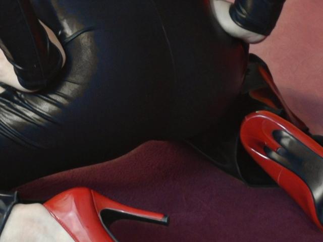 Los pantalones de cuero negro, los guantes negros y los tacones rojos de AshlyChloe están en una pose ajustada, sexy y provocativa sobre una alfombra roja. Sus brillantes tacones rojos están prominentemente exhibidos..