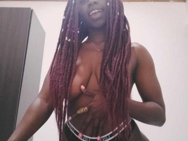 AndyCorrea69, mit dunkler Haut, langen roten Zöpfen, oben ohne, wobei sie ihre Brüste mit einer Hand bedeckt, trägt schwarze und weiße Victoria's Secret Unterwäsche und lächelt in einem Raum..