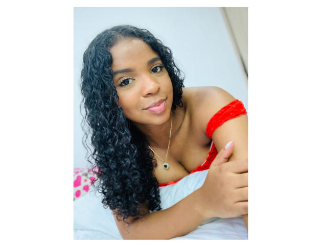 LisaMendez, con cabello negro rizado, una blusa roja con un hombro descubierto que muestra el escote, sonriendo, haciendo un gesto de pulgar hacia arriba, llevando un collar dorado con un colgante negro, sobre una cama blanca..