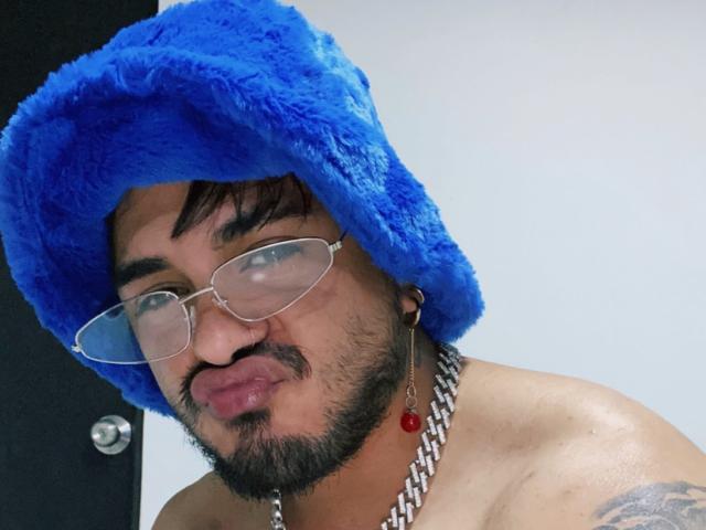 VisiI, con un sombrero azul y peludo, gafas y un collar de cadena, está haciendo pucheros y tiene un pequeño tatuaje en el hombro derecho. VisiI tiene barba y cabello negro corto..