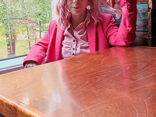 MissAstravert, avec ses cheveux roses, portant une veste rose et une chemise rose à boutons, est assise à une table en bois, levant sa main droite, et montrant ses boucles d'oreilles roses..