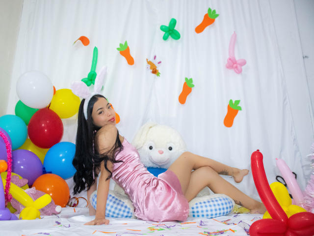 MisSofia vestida com um vestido de cetim rosa, cabelos longos e pretos, deitada em um travesseiro azul quadriculado, cercada por balões coloridos, decorações de papel de cenoura e borboleta, um coelho de pelúcia branco, um brinquedo sexual vermelho..