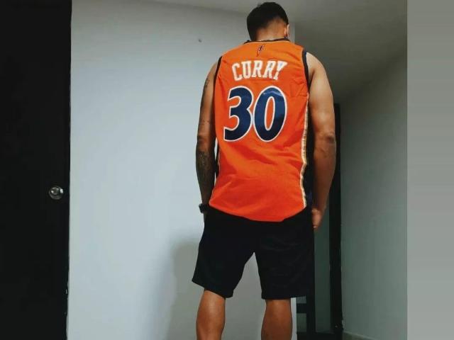 VisiI está de espaldas a la cámara, vistiendo una camiseta naranja de baloncesto con la inscripción "CURRY 30" en blanco y azul, y pantalones cortos negros, en una habitación sencilla..