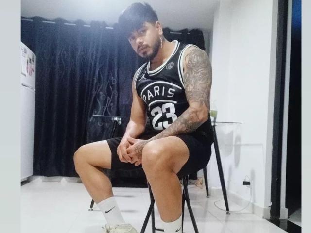 Een persoon met tatoeages op de armen, een zwart basketbalshirt met de tekst "Paris 23", zwarte korte broek, witte sokken en sneakers, zittend op een stoel, naar beneden kijkend, met de handen op de knieën, in een witte kamer..