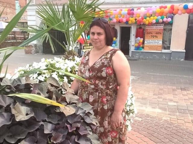 HayleyLady, een wat mollige vrouw met kort bruin haar, gekleed in een bruine jurk met bloemenprint, staat voor witte bloemen en paarse bladeren, met kleurrijke ballonnen op de achtergrond..
