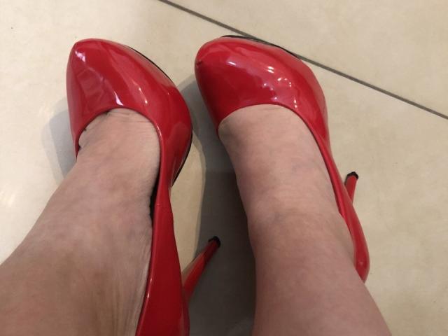 LadyMiller's pale legs wearing shiny red high heels with pointed toes and black heels, standing on a beige tiled floor.