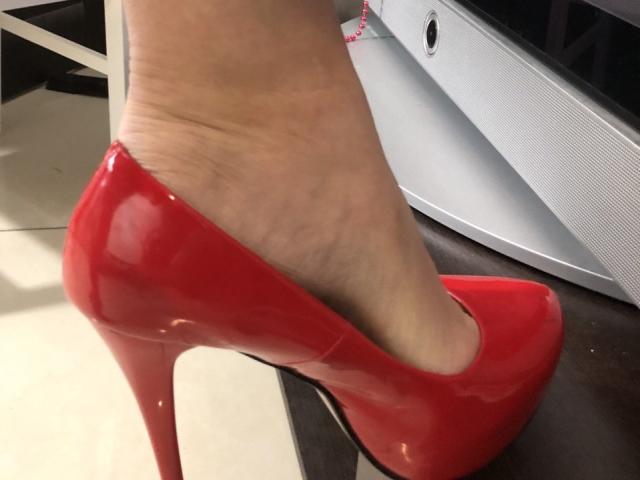 LadyMiller's foot in shiny red high heels, showing her smooth, pale skin and the stiletto heel's reflection. Her ankle's slightly visible, with a pink bracelet on her wrist.