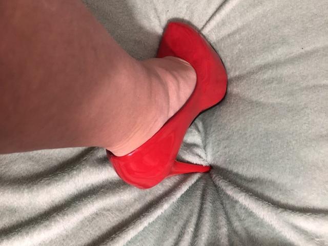 LadyMiller's foot in a bright red, high-heel shoe, showing her pale skin and slightly wrinkled leg, resting on a soft, light gray, textured surface.