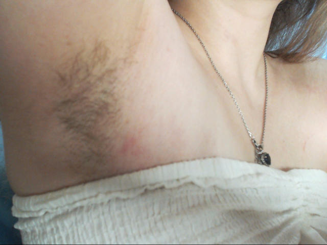 AliceReeve's armpit with dark, coarse hair, slightly red skin, and a small blemish, wearing a white, strapless top and a silver necklace with a small black pendant.