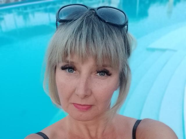 VAlica heeft kort blond haar, een zwarte zonnebril op haar hoofd, blauwe ogen, zwarte bandjes, roze lippenstift en een lichte glimlach. Op de achtergrond is zwembadwater te zien. Haar borsten zijn niet zichtbaar..