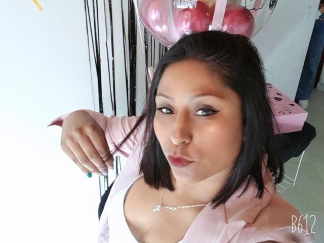 RoxyLatin, met een medium bruine huid, zwart steil haar en roze lippenstift, draagt een roze jas en een witte top, waarbij haar kleine borsten en ketting zichtbaar zijn, en ze maakt een ondeugende grimas..