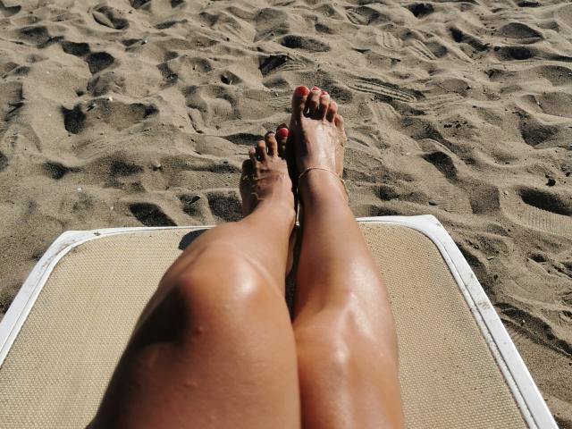 De glanzende, bruine benen van PassionX, met rode nagellak, liggen op een strandlaken, met zand overal om haar heen. Ze draagt een gouden enkelband, en haar voeten zijn licht bevuild door het zand..